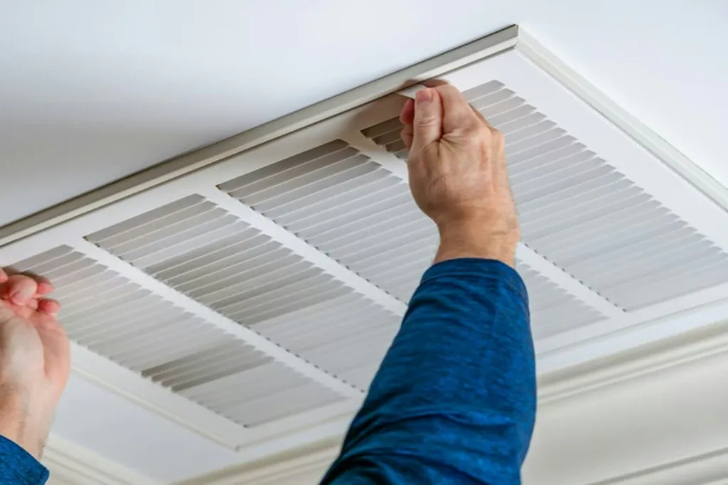 Hands lowering a ceiling return air grille to reach the filter during a scheduled maintenance visit