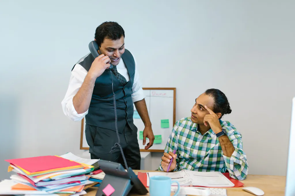 A busy shop office, one person on the desk phone while a coworker sits at a desk piled with job folders, a hand on their head